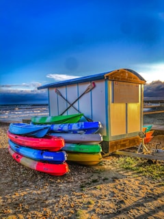 Several colorful kayaks are stacked next to a small wooden hut on a pebbled beach, with the sea and a dramatic blue sky with some clouds in the background. The hut has a sign indicating kayak rentals.