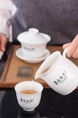 A white ceramic teapot pours light brown tea into a matching white teacup with a lid on the table nearby. The set rests on a bamboo tray, and a person is visible in the background.