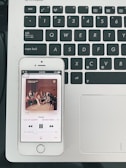 A smartphone displaying a music player app, showing the song 'Home' by SEVENTEEN, is placed on a laptop keyboard. The phone's screen shows album artwork featuring a group of people seated in a room. The laptop keyboard is black with white lettering, and the laptop's surface is silver.