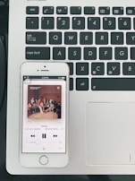 A smartphone displaying a music player app, showing the song 'Home' by SEVENTEEN, is placed on a laptop keyboard. The phone's screen shows album artwork featuring a group of people seated in a room. The laptop keyboard is black with white lettering, and the laptop's surface is silver.