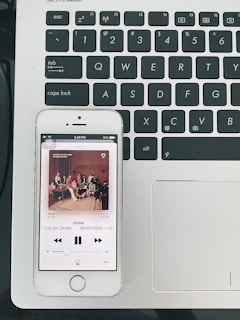 A smartphone displaying a music player app, showing the song 'Home' by SEVENTEEN, is placed on a laptop keyboard. The phone's screen shows album artwork featuring a group of people seated in a room. The laptop keyboard is black with white lettering, and the laptop's surface is silver.