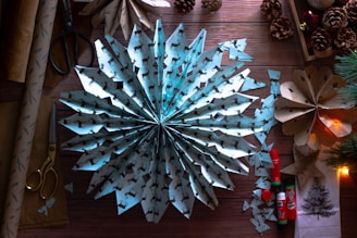 A neatly arranged workspace features a large, intricately folded paper snowflake with reindeer patterns. Surrounding it are various crafting materials including scissors, glue sticks, pine cones, and rolls of wrapping paper. A warm, cozy atmosphere is accentuated by subtle lighting, and additional decorations like a small branch of pine needles add to the festive feel.