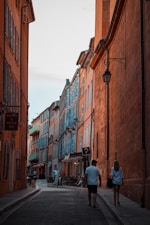 A narrow, picturesque street flanked by tall, colorful buildings with shutters, showcasing a mix of oranges and blues. The street is populated with pedestrians enjoying a leisurely walk. A dimly lit atmosphere with warm, ambient lighting emanating from small shops and cafes. A vintage-style street lamp enhances the quaint, cozy feel of the scene.