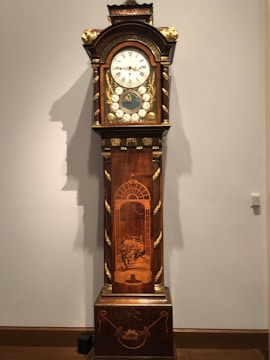 An ornate grandfather clock with a wooden casing featuring intricate carvings and gold detailing. The clock face has roman numerals and additional dials along with decorative elements. The lower section of the clock displays a detailed inlay scene with figures in a classical setting, framed by an arch.