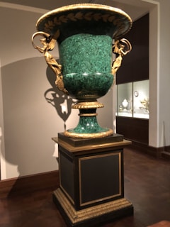 A large decorative urn made of green marble with intricate gold embellishments, including sculpted figures and leaf motifs. It is mounted on a pedestal in a museum-like setting with other decorative artifacts displayed in the background.