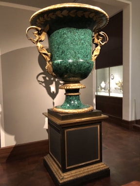 A large decorative urn made of green marble with intricate gold embellishments, including sculpted figures and leaf motifs. It is mounted on a pedestal in a museum-like setting with other decorative artifacts displayed in the background.