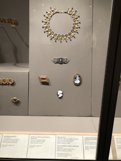 A display case contains a collection of elegant jewelry pieces, including a large gold and gemstone necklace, brooches, and decorative belt buckles. The jewelry is arranged neatly on a gray background with accompanying informational labels beneath each piece.