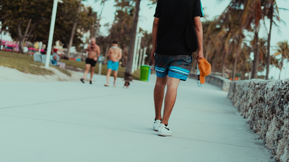 Hombre con camiseta negra y pantalones cortos de mezclilla azul caminando sobre piso de concreto blanco durante el día