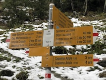 A multi-directional hiking signpost in a snowy, mountainous area, surrounded by evergreen trees. The sign provides various route options with estimated travel times to destinations like Alpe Garzott, Cap. Motterascio, Crap la Crusch, Larecc, Sentiero Luzzone, Diga Luzzone, Campo Blenio, and Olivone. The elevation is indicated as 1635 meters.