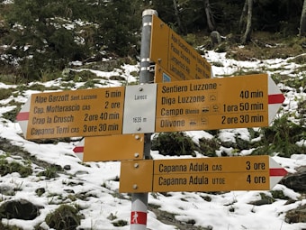A multi-directional hiking signpost in a snowy, mountainous area, surrounded by evergreen trees. The sign provides various route options with estimated travel times to destinations like Alpe Garzott, Cap. Motterascio, Crap la Crusch, Larecc, Sentiero Luzzone, Diga Luzzone, Campo Blenio, and Olivone. The elevation is indicated as 1635 meters.