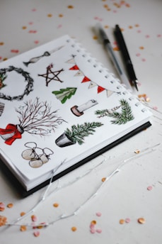 A sketchbook page filled with various festive illustrations such as a wreath, stars, candles, Christmas trees, and branches with decorations, accompanied by two pens placed beside it. The background features scattered confetti in pink and orange hues.
