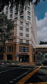 A tall building with a modern design, featuring a hospital entrance on the ground floor. The street intersection in front has traffic markings and a pedestrian crossing. Trees and parked vehicles are present, contributing to an urban environment.