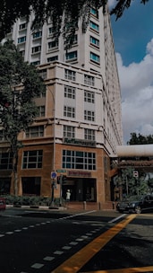 A tall building with a modern design, featuring a hospital entrance on the ground floor. The street intersection in front has traffic markings and a pedestrian crossing. Trees and parked vehicles are present, contributing to an urban environment.