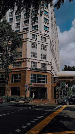 A tall building with a modern design, featuring a hospital entrance on the ground floor. The street intersection in front has traffic markings and a pedestrian crossing. Trees and parked vehicles are present, contributing to an urban environment.