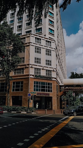 A tall building with a modern design, featuring a hospital entrance on the ground floor. The street intersection in front has traffic markings and a pedestrian crossing. Trees and parked vehicles are present, contributing to an urban environment.