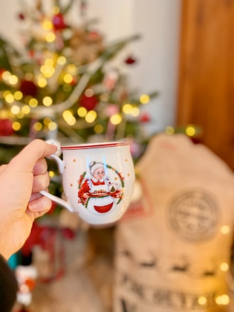 A hand holding a festive mug with an image of Mrs. Claus in front of a decorated Christmas tree with colorful ornaments and lights. In the background, a large sack and other holiday decorations are visible, creating a warm, festive atmosphere.
