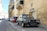 A classic Volkswagen car is parked on a narrow European street lined with stone buildings. The street is slightly inclined, and several other cars are parked along the curb. The buildings have a rustic, historic appearance with balconies and shuttered windows.