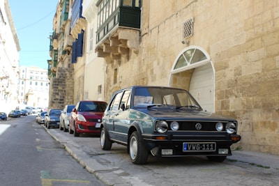 A classic Volkswagen car is parked on a narrow European street lined with stone buildings. The street is slightly inclined, and several other cars are parked along the curb. The buildings have a rustic, historic appearance with balconies and shuttered windows.