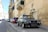 A classic Volkswagen car is parked on a narrow European street lined with stone buildings. The street is slightly inclined, and several other cars are parked along the curb. The buildings have a rustic, historic appearance with balconies and shuttered windows.
