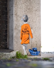 A stencil-style street art depicts a child with grey hair facing away, wearing an orange raincoat with the words 'Sea Rescue is not a crime' printed on the back. The child pulls a blue toy boat on a string. The artwork is painted on a textured concrete wall next to a darker brick wall, with grass and pavement visible below.