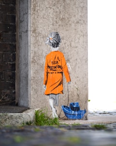 A stencil-style street art depicts a child with grey hair facing away, wearing an orange raincoat with the words 'Sea Rescue is not a crime' printed on the back. The child pulls a blue toy boat on a string. The artwork is painted on a textured concrete wall next to a darker brick wall, with grass and pavement visible below.