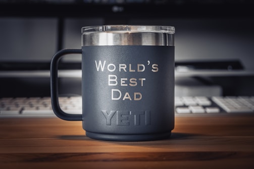 A matte black mug with the words 'World's Best Dad' printed in white sits on a wooden desk. The brand name 'YETI' is embossed near the bottom of the mug. In the background, there is a keyboard and a partially visible monitor, creating a workspace environment.