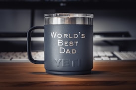 A matte black mug with the words 'World's Best Dad' printed in white sits on a wooden desk. The brand name 'YETI' is embossed near the bottom of the mug. In the background, there is a keyboard and a partially visible monitor, creating a workspace environment.