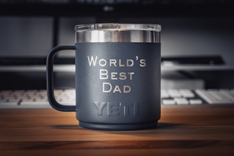 A matte black mug with the words 'World's Best Dad' printed in white sits on a wooden desk. The brand name 'YETI' is embossed near the bottom of the mug. In the background, there is a keyboard and a partially visible monitor, creating a workspace environment.