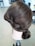 A mannequin head with neatly styled brown hair in an elegant updo. The hairstyle features intricate twists and curls, and the mannequin is positioned on a metal stand in a room with office furniture visible in the background.
