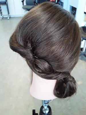 A mannequin head with neatly styled brown hair in an elegant updo. The hairstyle features intricate twists and curls, and the mannequin is positioned on a metal stand in a room with office furniture visible in the background.