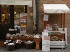 A small outdoor market stall and shop entrance with a variety of products displayed. The stall features wicker baskets filled with packaged goods, jars, and bottles. Several signs and price tags are visible, along with a banner above the entrance. The setting appears rustic with a brick wall backdrop.