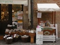 A small outdoor market stall and shop entrance with a variety of products displayed. The stall features wicker baskets filled with packaged goods, jars, and bottles. Several signs and price tags are visible, along with a banner above the entrance. The setting appears rustic with a brick wall backdrop.