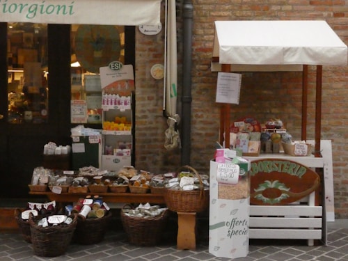 A small outdoor market stall and shop entrance with a variety of products displayed. The stall features wicker baskets filled with packaged goods, jars, and bottles. Several signs and price tags are visible, along with a banner above the entrance. The setting appears rustic with a brick wall backdrop.
