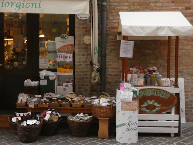 A small outdoor market stall and shop entrance with a variety of products displayed. The stall features wicker baskets filled with packaged goods, jars, and bottles. Several signs and price tags are visible, along with a banner above the entrance. The setting appears rustic with a brick wall backdrop.