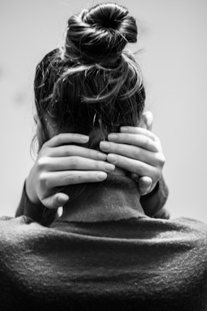 A black-and-white photo of someone with their back to the camera. The person has long hair tied in a bun and is holding their neck with both hands. The image is focused on the upper back, neck, and hands.
