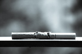 A cigarette resting on a flat surface with its paper partially torn, exposing the tobacco inside. The image is black and white, giving it a stark and dramatic appearance. The background is blurred, highlighting the focus on the cigarette.