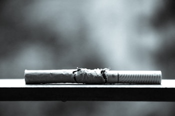 A cigarette resting on a flat surface with its paper partially torn, exposing the tobacco inside. The image is black and white, giving it a stark and dramatic appearance. The background is blurred, highlighting the focus on the cigarette.