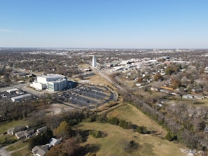 Aerial view of a suburban area featuring a mix of residential, commercial, and industrial buildings. There is a large white building with an adjacent parking lot, a tall white structure resembling a tower, and sprawling residential neighborhoods with scattered trees and open spaces. Roads and railway tracks can be seen crisscrossing the area, and the landscape is predominantly flat.