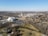 Aerial view of a suburban area featuring a mix of residential, commercial, and industrial buildings. There is a large white building with an adjacent parking lot, a tall white structure resembling a tower, and sprawling residential neighborhoods with scattered trees and open spaces. Roads and railway tracks can be seen crisscrossing the area, and the landscape is predominantly flat.