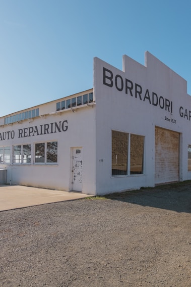 A vintage auto repair building with large lettering on the facade indicating 'Borradori Garage' and 'Auto Repairing'. The structure is painted white with a slightly weathered appearance. There are large windows on the side and peeling paint on the door, suggesting age and use.
