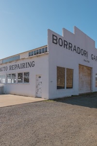 A vintage auto repair building with large lettering on the facade indicating 'Borradori Garage' and 'Auto Repairing'. The structure is painted white with a slightly weathered appearance. There are large windows on the side and peeling paint on the door, suggesting age and use.