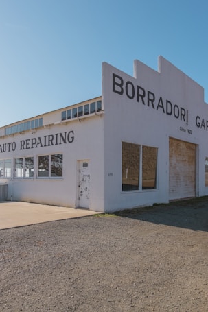 A vintage auto repair building with large lettering on the facade indicating 'Borradori Garage' and 'Auto Repairing'. The structure is painted white with a slightly weathered appearance. There are large windows on the side and peeling paint on the door, suggesting age and use.