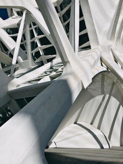Complex architectural structure with intertwining white beams and geometric patterns creating shadows. The design appears modern and abstract, emphasizing angles and lines.
