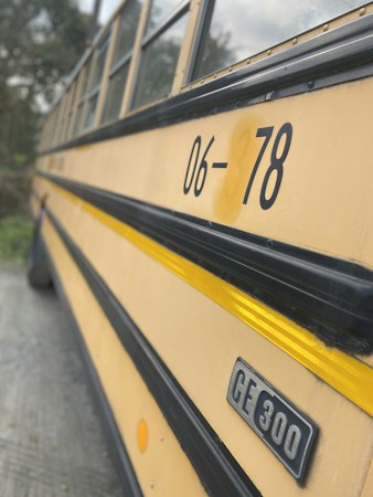 The image is a close-up of a yellow school bus with the number 06-378 and a CE 300 badge displayed prominently. The bus features a distinctive yellow color with a black horizontal stripe. The windows of the bus are visible at the top portion, along with surrounding greenery reflected on them.