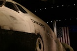 A close-up view of the space shuttle Discovery, prominently displaying its name on the side of the shuttle. The fuselage and windows are visible, with an American flag hanging in the background. The lighting is dim, suggesting an indoor setting, possibly a museum.