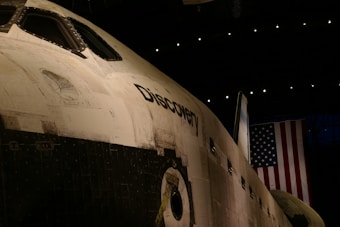 A close-up view of the space shuttle Discovery, prominently displaying its name on the side of the shuttle. The fuselage and windows are visible, with an American flag hanging in the background. The lighting is dim, suggesting an indoor setting, possibly a museum.