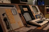 A vintage control room setup featuring old computer equipment, including large monitors, rotary dials, push buttons, and analog telephones. The panels are arranged with paper notes and diagrams attached, indicating a focus on monitoring and communication. The lighting is dim, creating a retro technological ambiance.