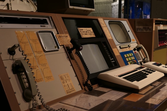 A vintage control room setup featuring old computer equipment, including large monitors, rotary dials, push buttons, and analog telephones. The panels are arranged with paper notes and diagrams attached, indicating a focus on monitoring and communication. The lighting is dim, creating a retro technological ambiance.