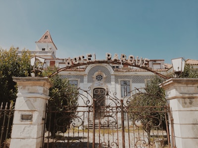 An old, ornate building with a sign reading 'Hotel Parque' at the entrance. The structure features decorative tiles, large windows, and a gated entrance with ironwork detailing. The building is surrounded by lush greenery, and the sky is clear.