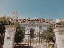 An old, ornate building with a sign reading 'Hotel Parque' at the entrance. The structure features decorative tiles, large windows, and a gated entrance with ironwork detailing. The building is surrounded by lush greenery, and the sky is clear.